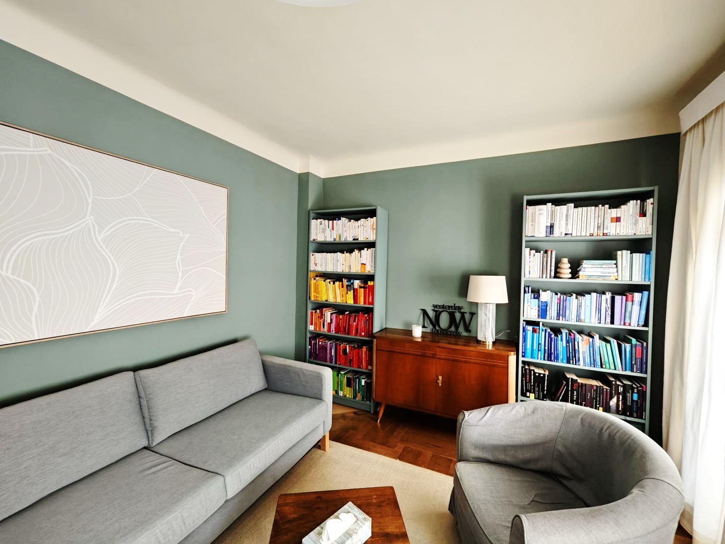 Therapieraum der Praxis: graues Sofa und Sessel, salbeigrüne Wände, Bücherregale und eine Holzkommode mit Leselampe.
